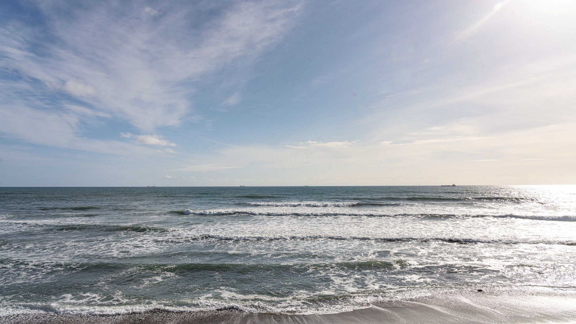 Une plage avec des vagues, un ciel dégagé et des navires à l’horizon.