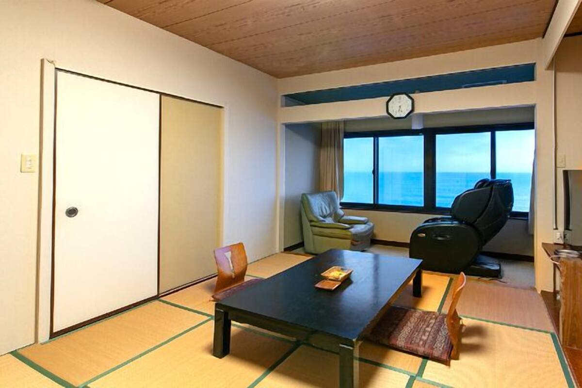 Una habitación con suelo de tatami, una mesa baja, una silla, un sofá, un televisor y una ventana con vistas.