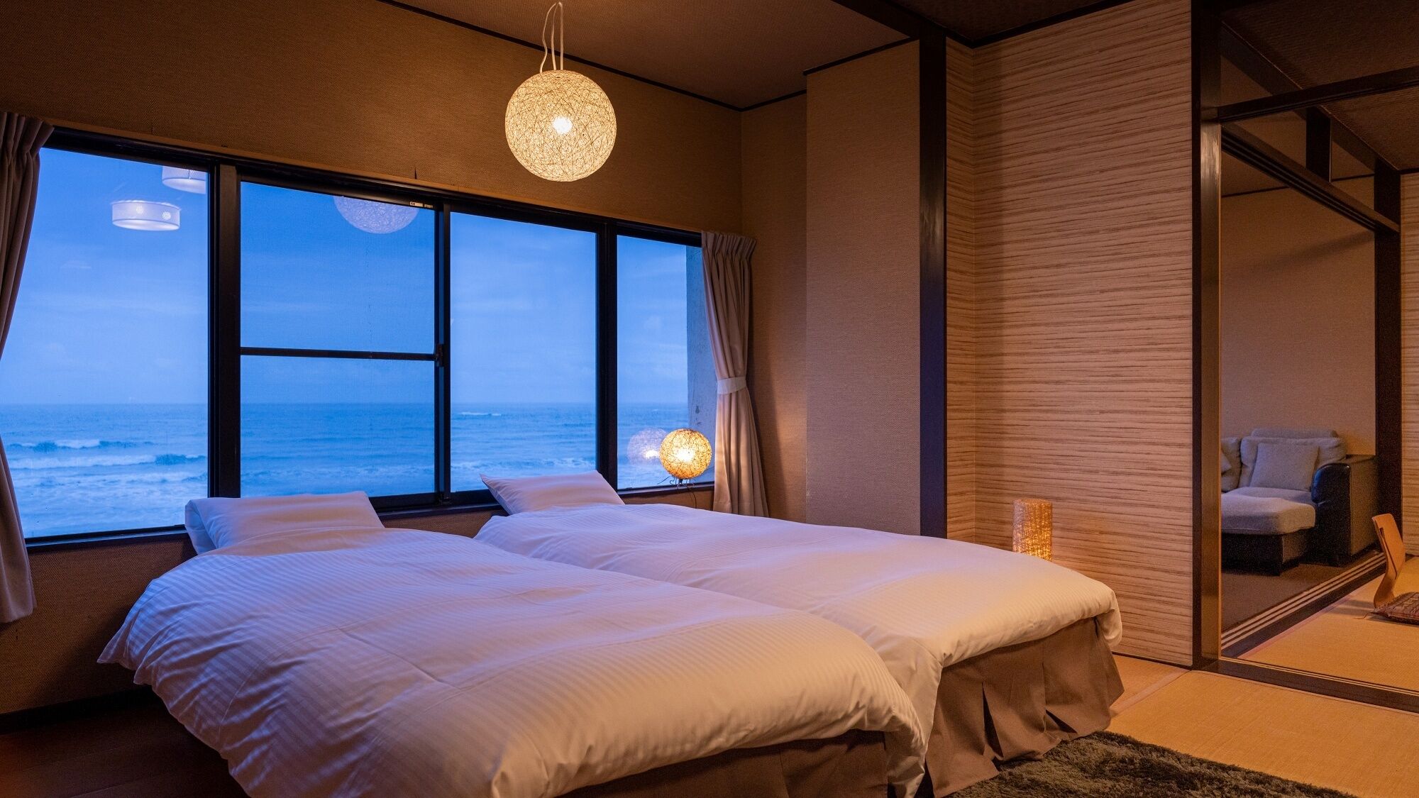 Une chambre avec deux lits, une grande fenêtre donnant sur l’océan, un luminaire suspendu et une porte coulissante qui mène à une autre pièce.