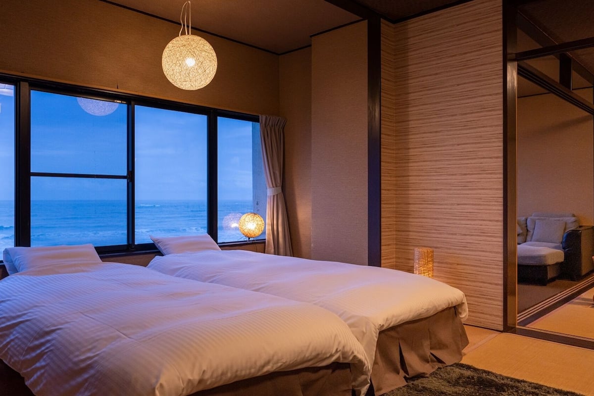 Une chambre avec deux lits, une grande fenêtre donnant sur l’océan, un luminaire suspendu et une porte coulissante qui mène à une autre pièce.