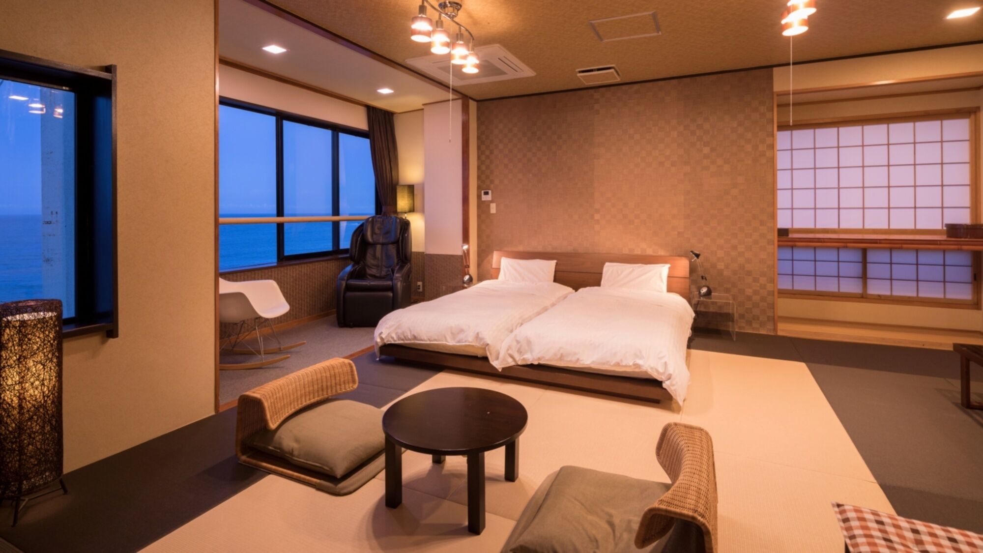 Une chambre d’hôtel comprenant un lit, deux chaises, une petite table, un fauteuil massant et de grandes fenêtres offrant une vue dégagée.