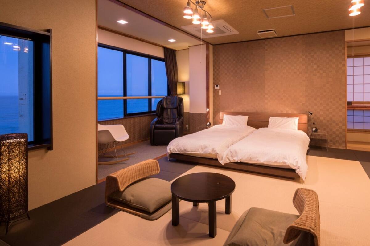 Habitación de hotel con cama, dos sillas, una mesita pequeña, un sillón de masaje y ventanas amplias con vistas.
