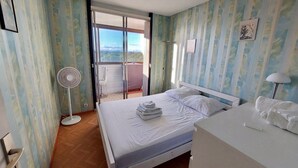 A bedroom with a bed, a bedside table, a fan, and a view of the outdoors. - 2-room apartment with balcony and parking, 250m from the beach (La Grande-Motte)