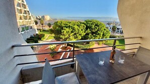 A balcony with a table and chairs, overlooking a landscaped area with trees and a distant view of a cityscape. - 2-room apartment with balcony and parking, 250m from the beach (La Grande-Motte)