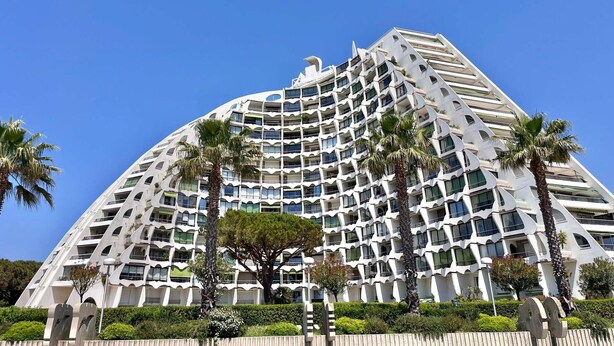 A modern hotel with a unique curved architecture, multiple balconies, and palm trees in the foreground. - 2-room apartment with balcony and parking, 250m from the beach (La Grande-Motte)