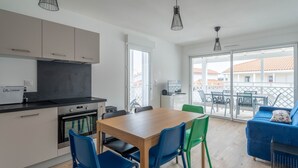 Una cocina moderna con armarios blancos, encimera negra y horno empotrado. Se observa una zona de comedor con una mesa de madera y sillas azules y verdes. Un sofá azul es visible al fondo, y la habitación tiene ventanas amplias.