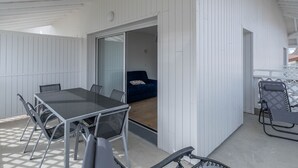 Un espacio exterior techado con una mesa y sillas, un ventanal corredero que da a una habitación con un sofá azul, y un balcón con una silla.