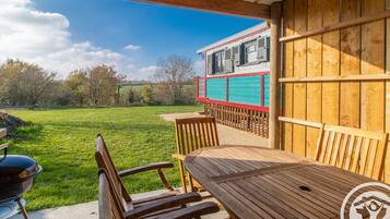 Une terrasse couverte avec une table et des chaises en bois, un barbecue et une vue sur une maison à la façade rouge et blanche.