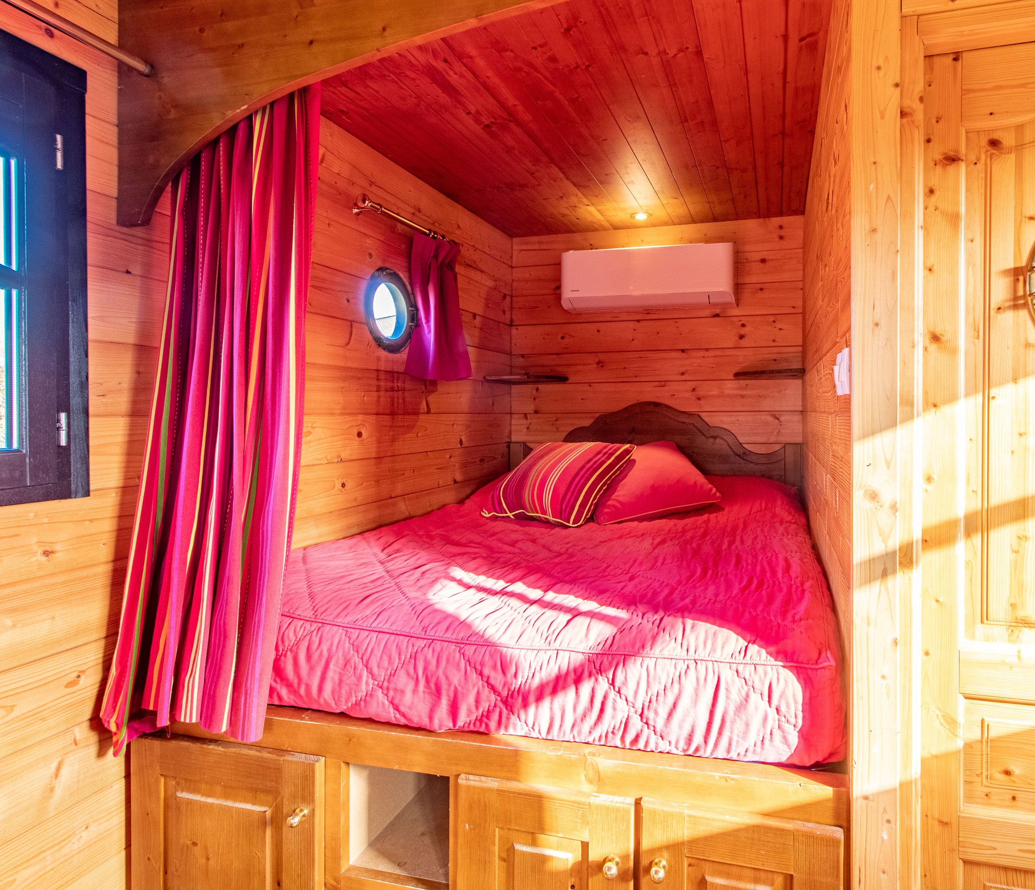 A wooden cabin room with a bed, a window with curtains, and a circular mirror on the wall.