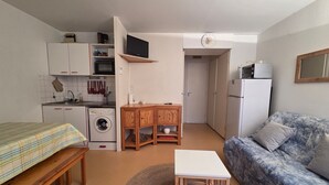Un espace de vie compact comprenant un coin cuisine, un lave-linge, un canapé, une petite table et un téléviseur fixé au mur.