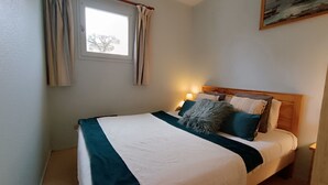 Un lit bien fait, recouvert d’une couvre-lit blanche et turquoise, avec une tête de lit en bois et une table de chevet sur laquelle se trouve une lampe.
