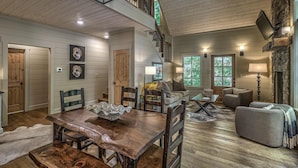 A spacious living room with a wooden dining table, a sofa, and a fireplace. - Cider Mill House overlooking private stream w/ firepit (Ellijay)