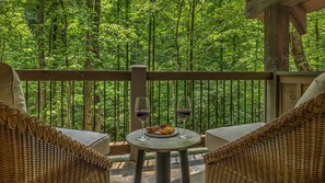 A balcony with wicker furniture, a table with a plate of snacks and two wine glasses, and a view of a dense forest. - Cider Mill House overlooking private stream w/ firepit (Ellijay)