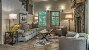 A cozy living room with a sofa, armchairs, a coffee table, and a fireplace. There are two windows with a view of greenery outside. - Cider Mill House overlooking private stream w/ firepit (Ellijay)