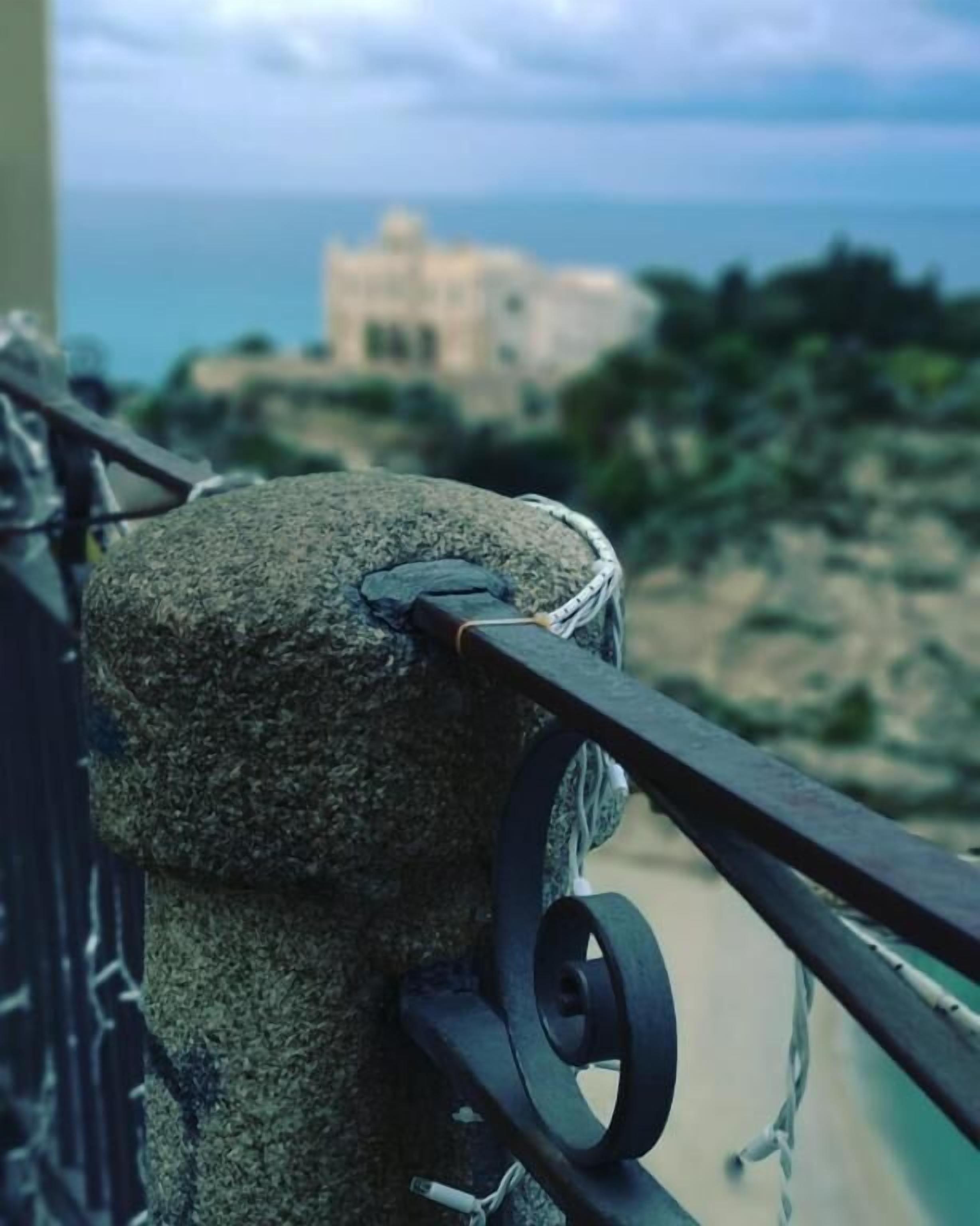 Un pilar de piedra con barandilla de metal, con vistas a una zona costera y edificios al fondo.