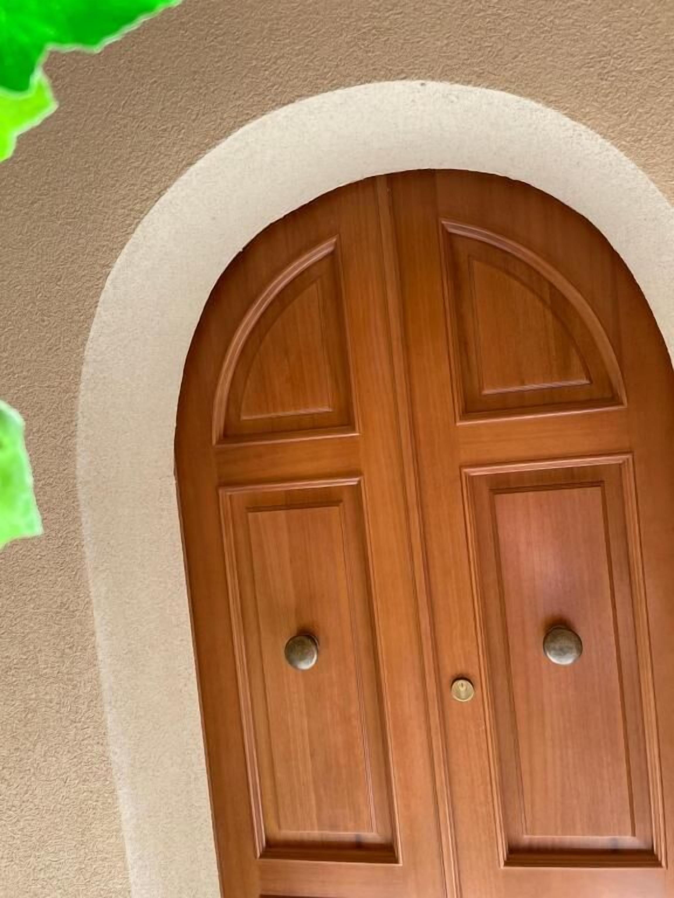 A wooden double door with a rounded arch above it, set in a beige wall.