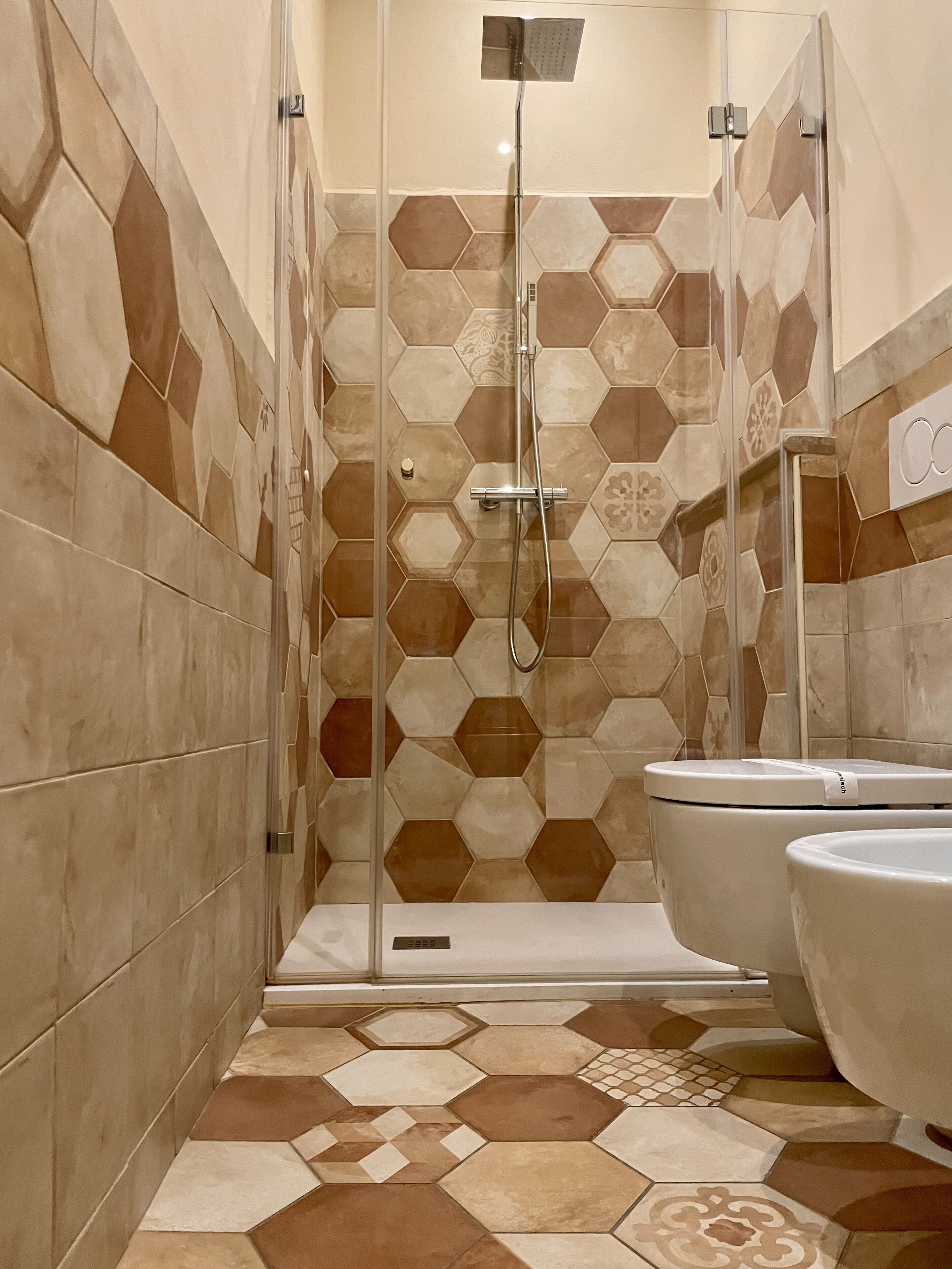 Un baño con baldosas de patrón hexagonal, ducha con mampara de vidrio y dos inodoros.