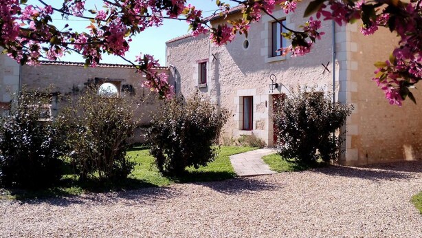 Un bâtiment rustique, avec un chemin de gravier menant à une porte, entouré d’arbustes en fleurs et surplombé d’un ciel bleu clair.