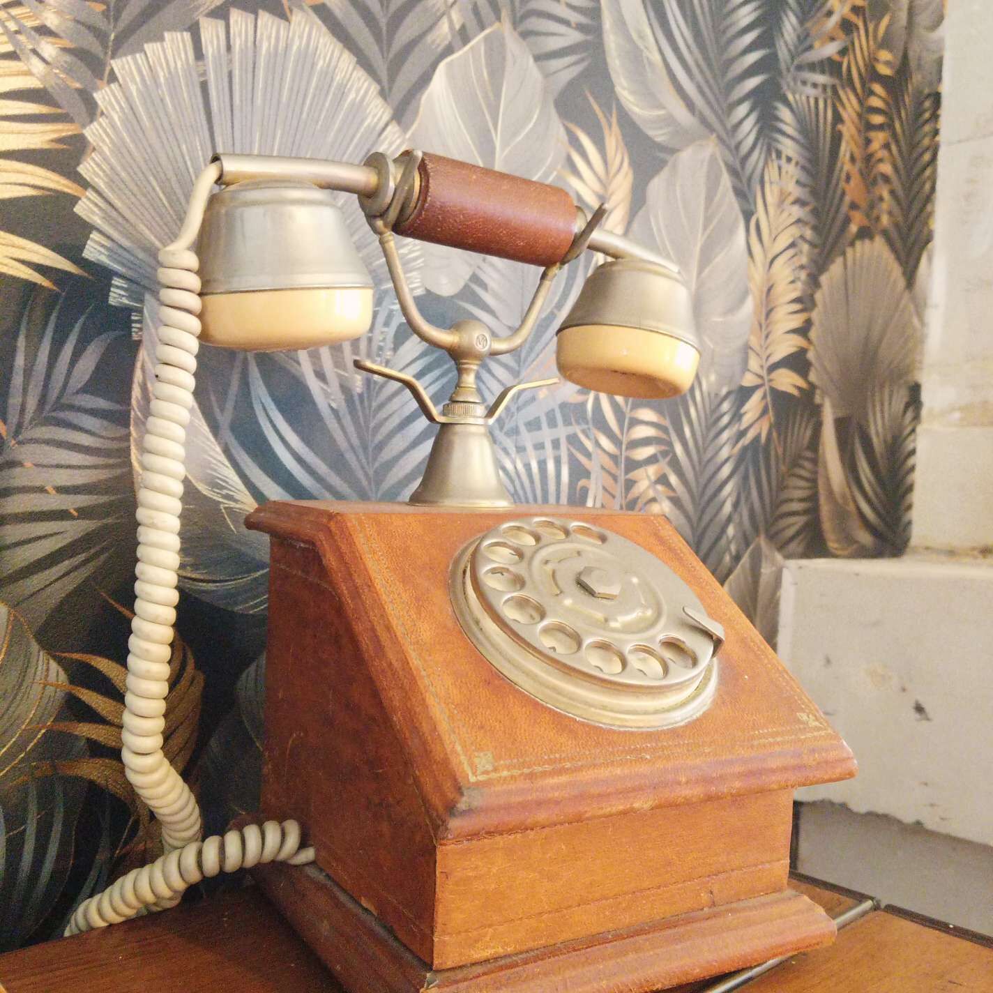 Un teléfono antiguo de disco con una base de madera, frente a un papel tapiz con motivos de hojas tropicales.