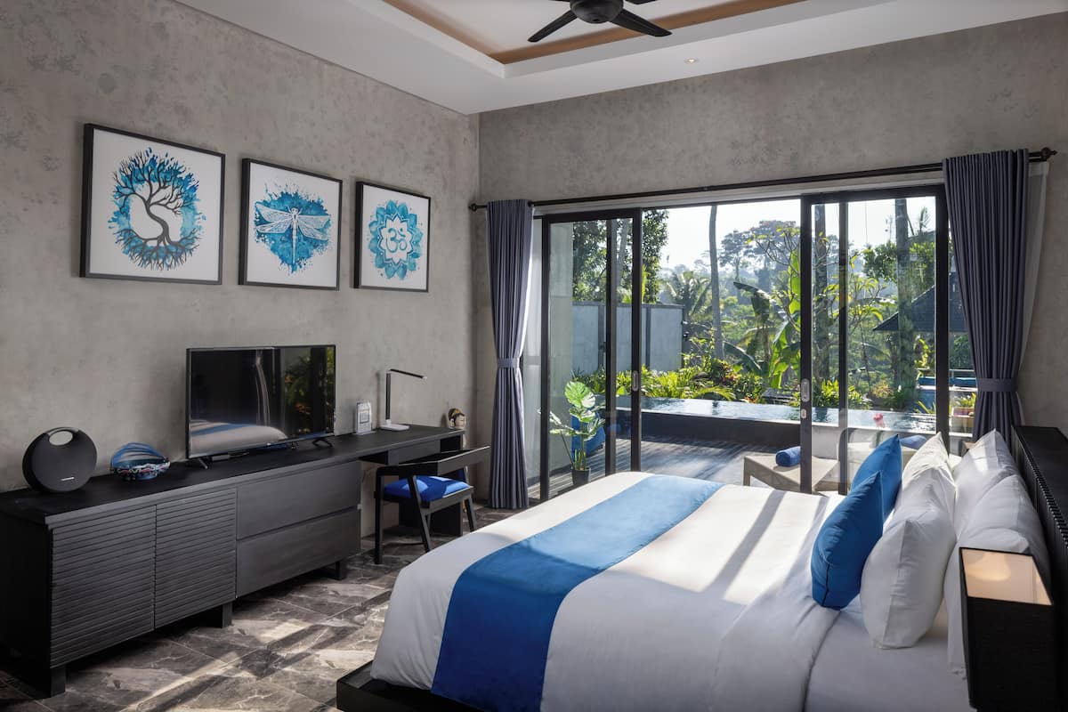 A modern hotel room with a large bed, a desk with a TV, and a balcony with a view of greenery.