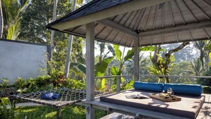 Un espace extérieur abrité avec des sièges, comprenant un hamac, un banc rembourré avec un plateau de nourriture, et une végétation tropicale luxuriante en arrière-plan.