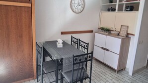 A compact kitchen with a dining table, chairs, a clock, and a white cabinet with shelves.