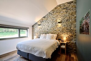 Une chambre avec un mur en pierre, un grand lit, une table de chevet et une fenêtre donnant sur un paysage verdoyant.