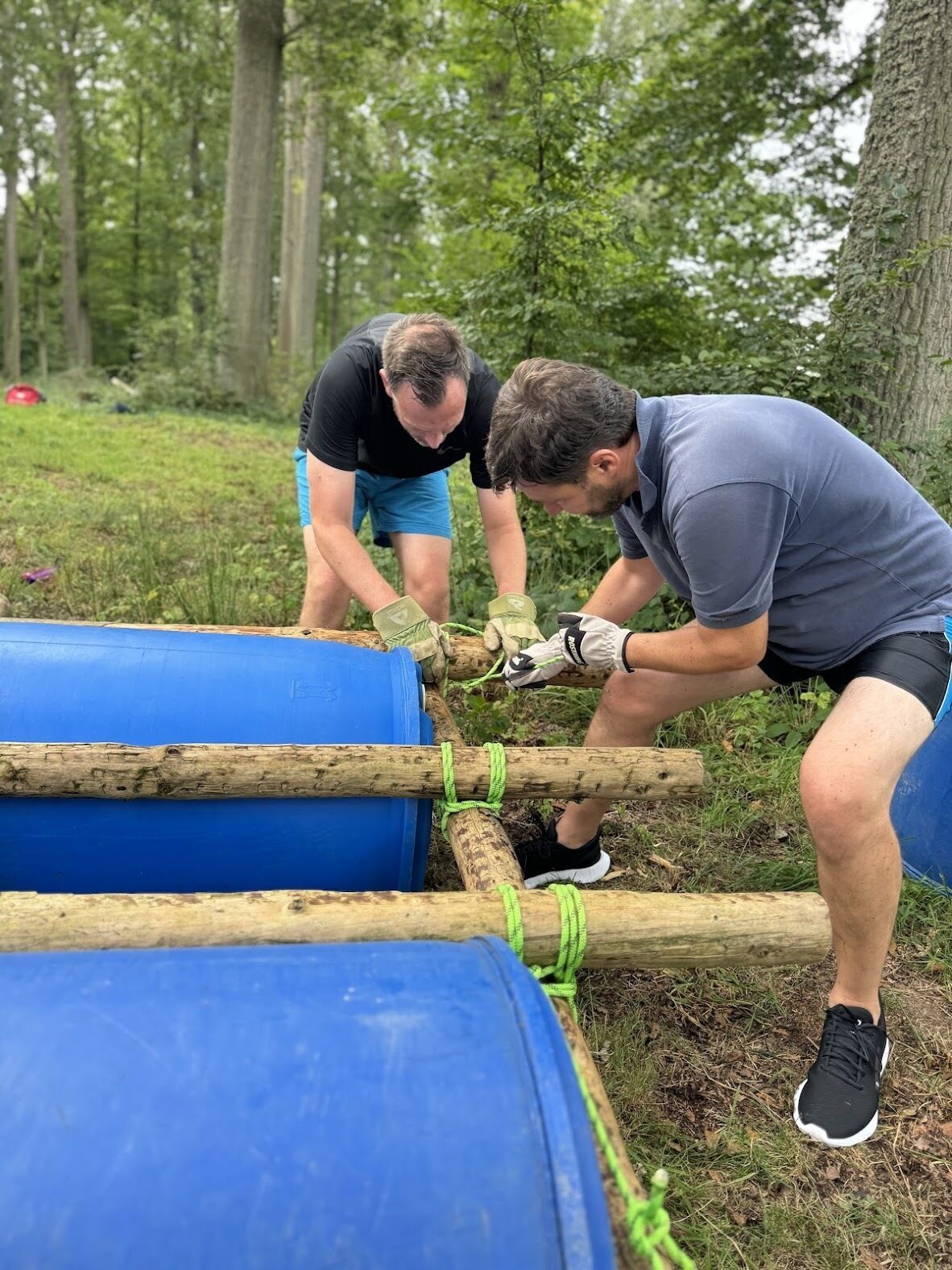 Twee personen bouwen een constructie met blauwe vaten en houten blokken in een bebost gebied.