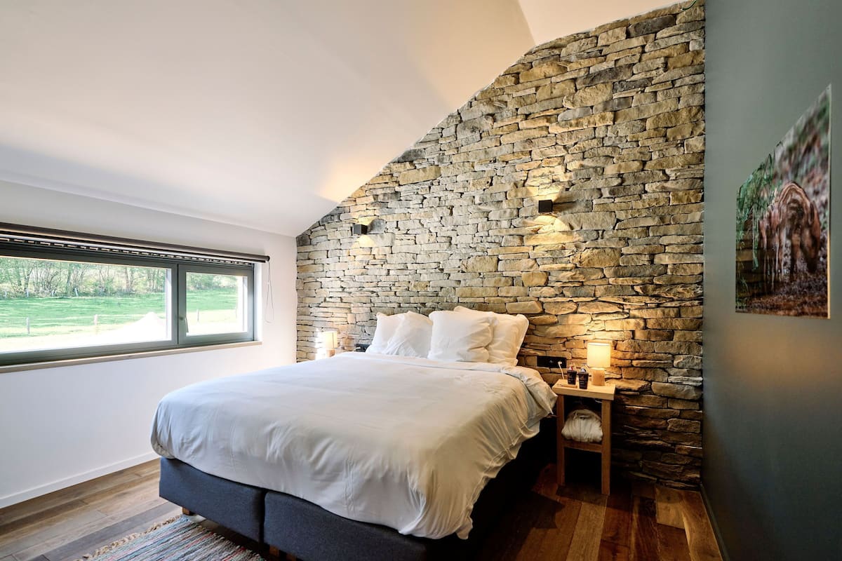 Une chambre avec un mur en pierre, un lit avec une literie blanche, une table de chevet avec une lampe et une fenêtre donnant sur un paysage verdoyant.