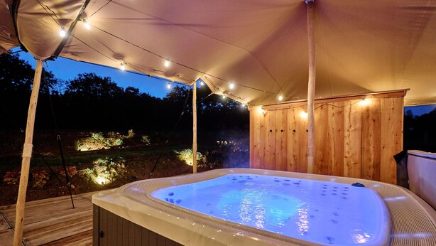 Un bain à remous installé sous une tonnelle décorée de guirlandes lumineuses, entouré de structures en bois et d’un jardin.