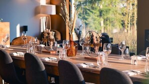 Un espace repas avec une longue table dressée pour un repas, entourée de chaises, et une vue sur les arbres à travers une grande fenêtre.