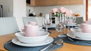 Una mesa de comedor dispuesta con tazas rosas, cuencos blancos y copas de vino, con un jarrón de flores al fondo.