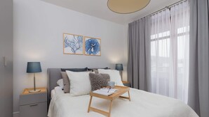 Un dormitorio moderno con una cama, mesitas de noche, una mesita de noche de madera con un libro y gafas, y una ventana con cortinas.