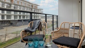 A balcony with wicker chairs, a table with glasses and a pitcher, and a view of a building and construction site. - Modern Apartment | Pool, Parking, Close to the Beach (Dziwnów)