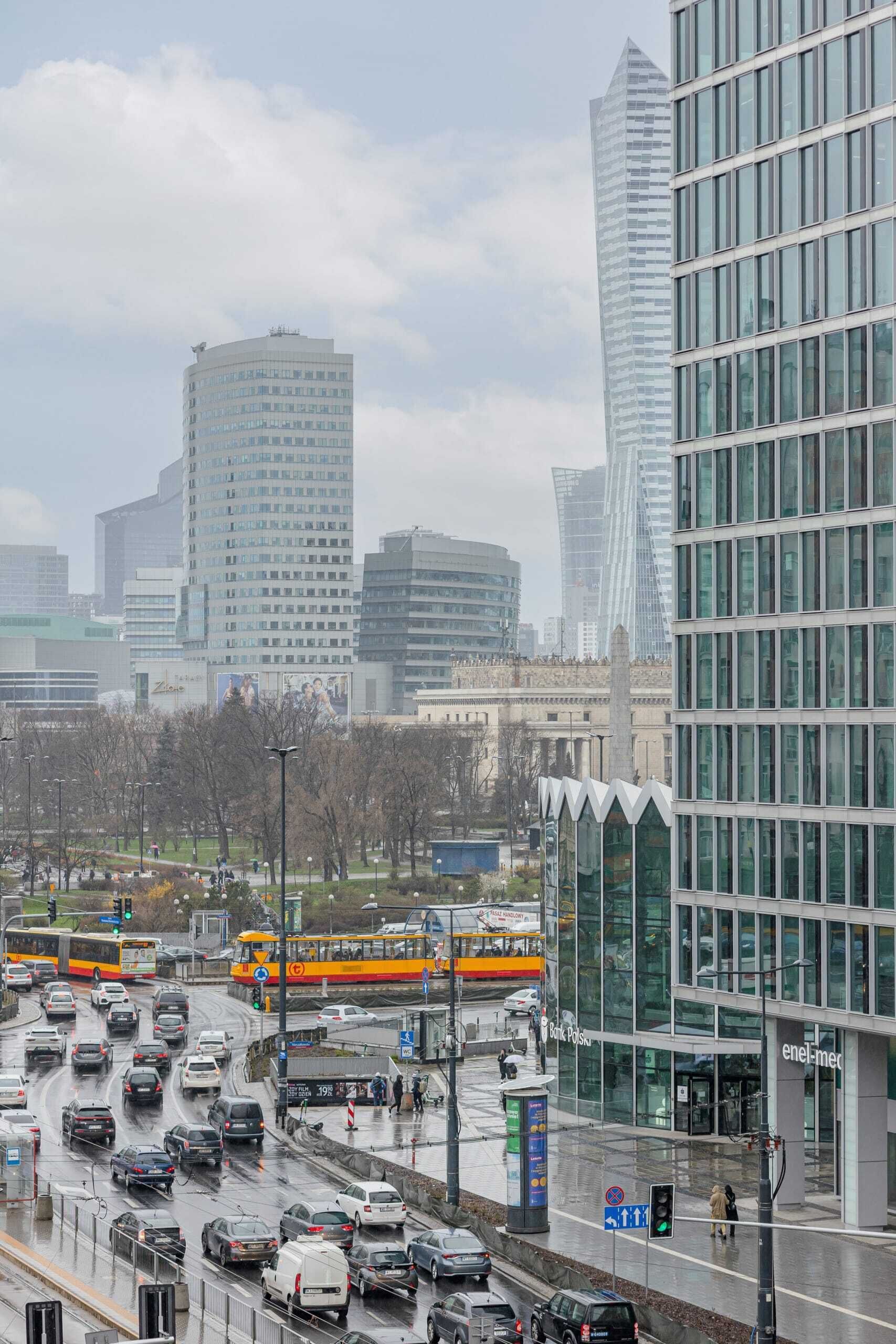 Un paysage urbain avec des bâtiments modernes, une rue animée où circulent des voitures et des autobus, et une ligne de tramway.