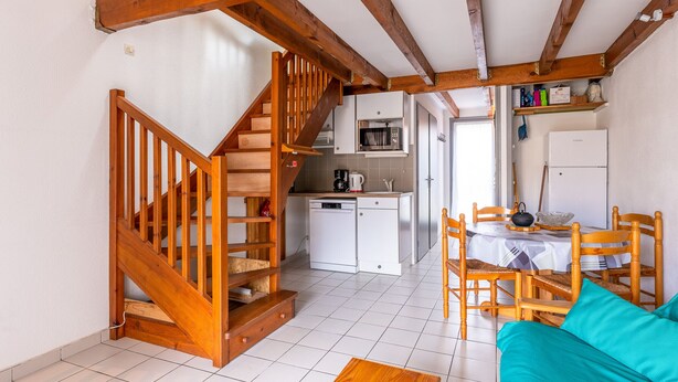 A cozy interior with a wooden staircase, a dining area with wooden chairs, a kitchen with white appliances, and a teal sofa. - Cozy Maisonette 300m from Beach in Biscarrosse – Sleeps 4, Terrace, Parking (Biscarrosse)