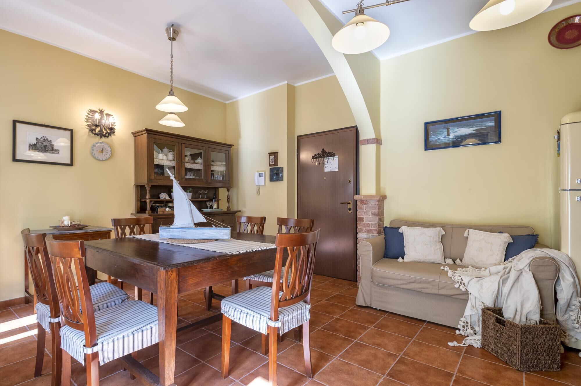 A dining area with a wooden table and chairs, a cabinet with glass doors, a sailboat model, and a sofa with pillows.