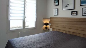 Un dormitorio con cama, ventana con persianas, lámpara de noche y cuadros en la pared.