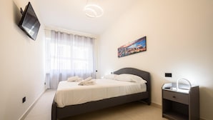 Een netjes ingerichte slaapkamer met een bed, nachtkastje, een aan de muur bevestigde televisie en een raam met gordijnen.