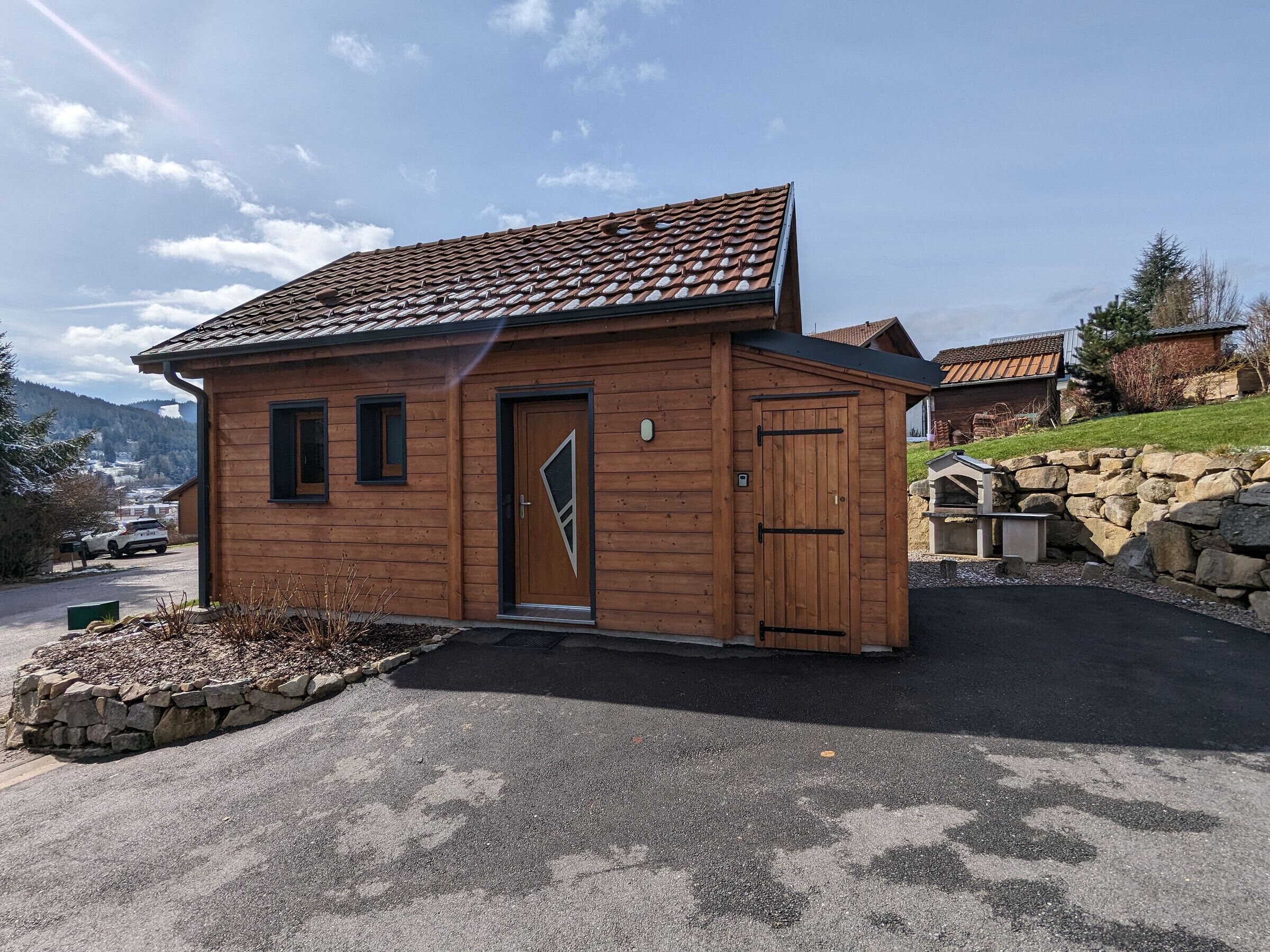 Una piccola baita di legno con tetto in tegole, una porta con finestra triangolare e un muro di pietra sullo sfondo.