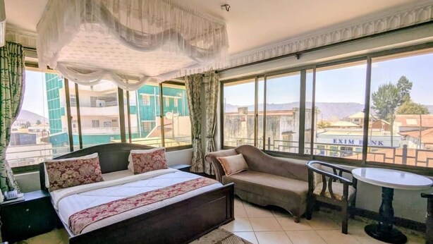 A hotel room with a bed, a sofa, a small table, and large windows offering a city view. - Alexander Hotel & Tours (Iringa)