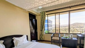 A hotel room with a large bed, a desk with a chair, a view of a cityscape through large windows, and a door. - Alexander Hotel & Tours (Iringa)