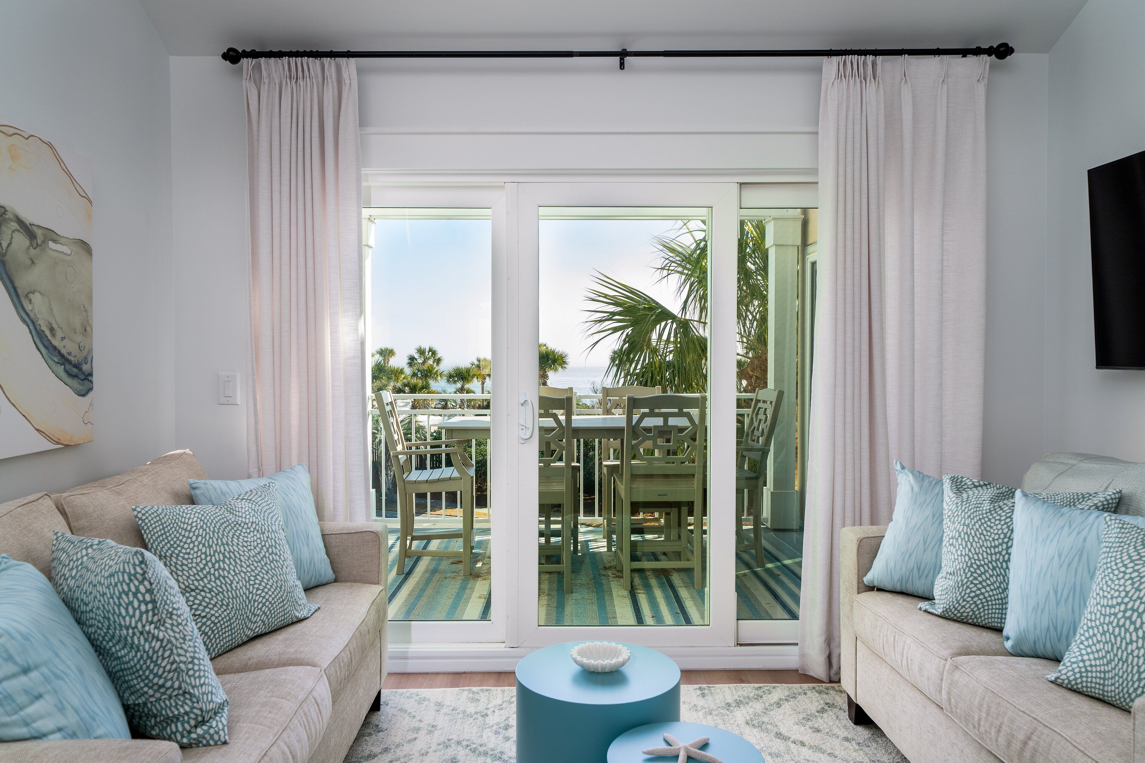 A living room with a beige sofa, blue patterned cushions, a round blue coffee table, and a glass door leading to an outdoor area with a table and chairs.