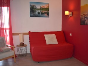 Um sofá vermelho com uma almofada branca, uma pequena mesa redonda com uma luminária e uma cadeira com um encosto. Uma pintura de uma paisagem montanhosa está na parede.