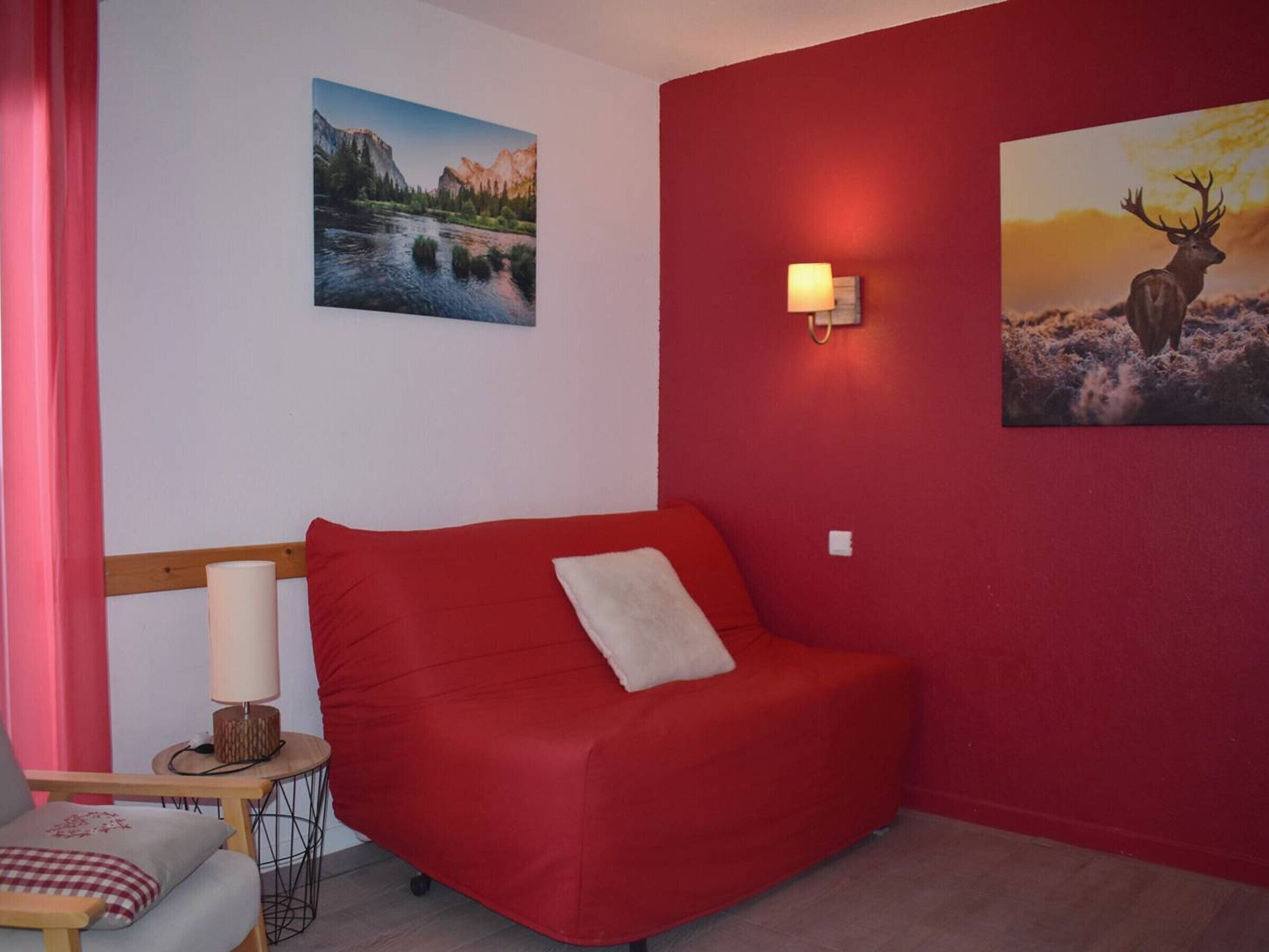 A red sofa with a white pillow, a small table with a lamp, and two framed pictures on the walls.