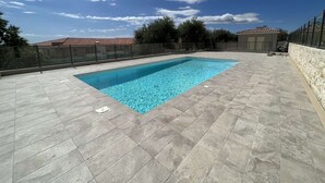 Una piscina rectangular con agua azul cristalina, rodeada por un solárium con baldosas y un muro de piedra.