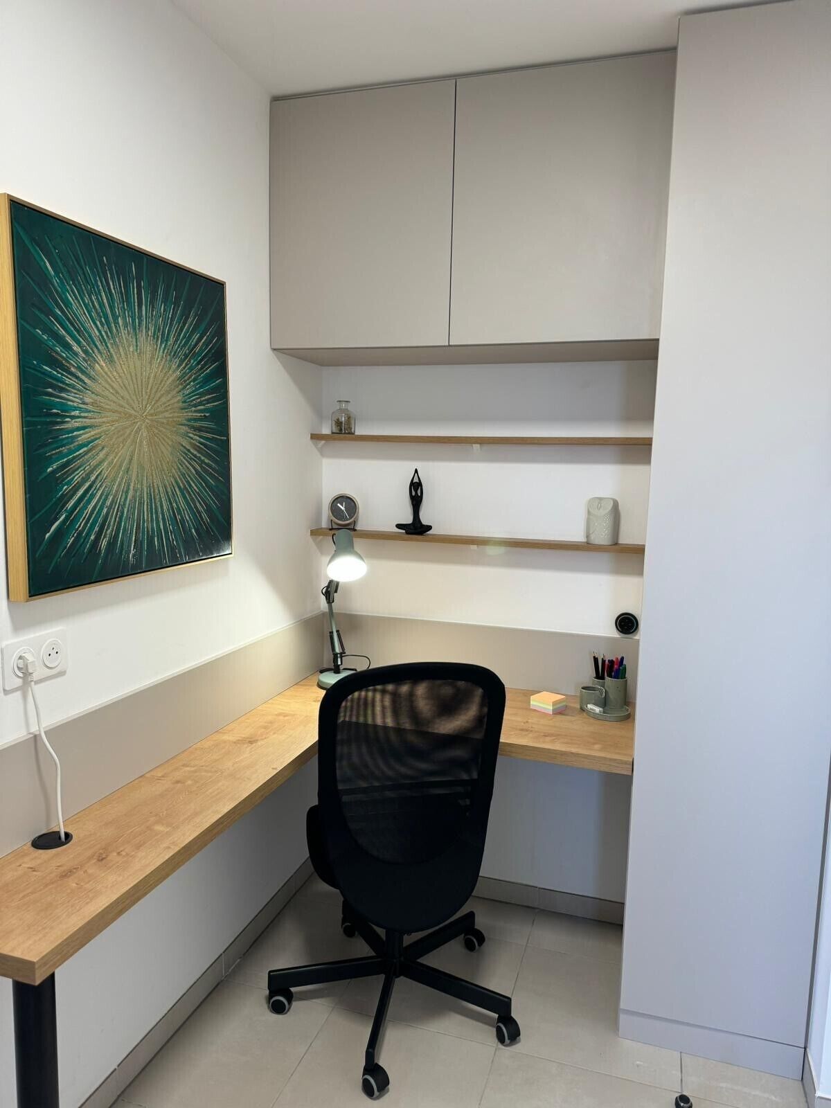 Un espace de bureau moderne avec un bureau en bois, une chaise de bureau noire, une lampe de bureau et un élément décoratif mural.
