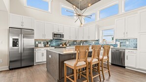 A modern kitchen with white cabinets, stainless steel appliances, a large island with chairs, and a view of the sky through the windows.
