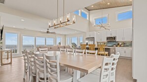A spacious dining area with a large wooden table and white chairs, a modern kitchen with white cabinets, and large windows offering a view of the ocean.
