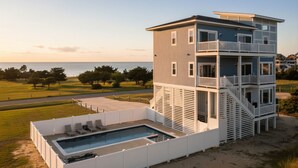 A multi-story building with a pool, located near a beach with trees and a clear sky.